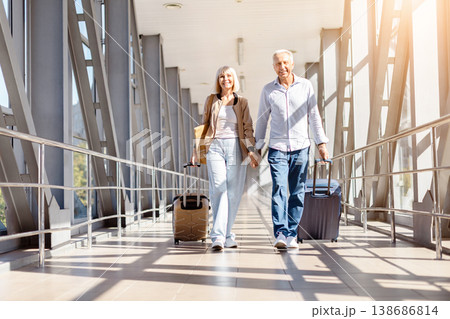 Mature couple walking together in airport terminal holding hands and pulling luggage Mature couple walking together in airport terminal holding hands and pulling luggage 138686814