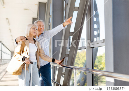 Mature man pointing through airport window while embracing wife with luggage 138686874