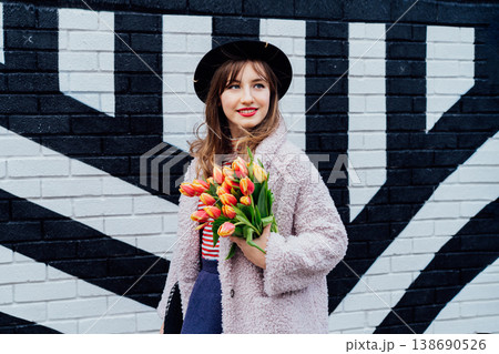 Portrait of happy. smiling young fashion woman holding bouquet of fresh tulip flowers on the painted brick wall background. Urban city street fashion. Spring mood. Happiness and positive vibes. Portrait of happy. smiling young fashion woman holding bouquet of fresh tulip flowers on the painted brick wall background. Urban city street fashion. Spring mood. Happiness and positive vibes. 138690526