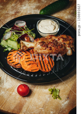 Roasted pork knuckle served with grilled sweet potato slices a fresh garden salad and creamy dipping sauce on a black plate. 138691027
