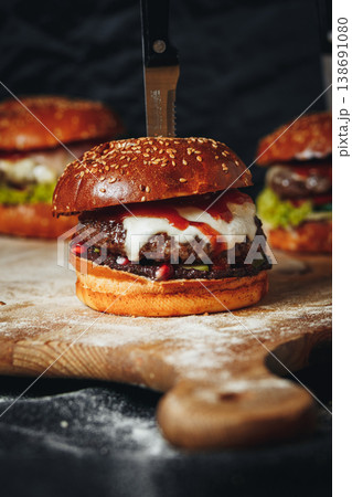 Gourmet beef burger with melted cheese and pomegranate seeds served on a rustic wooden board with a knife stuck through the bun. 138691080