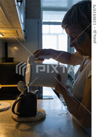 An elderly woman with dark circles under her eyes pours hot water from an electric kettle into teapot in her kitchen, enjoying her morning routine. Real people, life of an elderly person An elderly woman with dark circles under her eyes pours hot water from an electric kettle into teapot in her kitchen, enjoying her morning routine. Real people, life of an elderly person 138691979