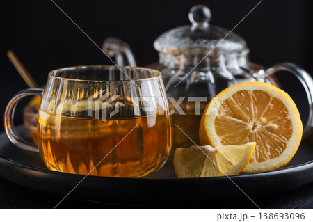 Glass teapot with amber tea, teacup with lemon slice, and bowl of honey with golden spoon on dark concrete background 138693096