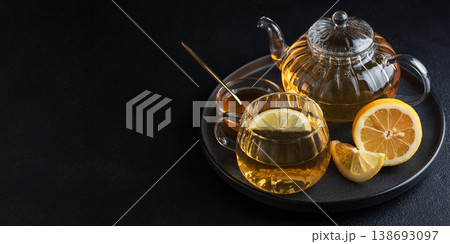 Glass teapot with amber tea, teacup with lemon slice, and bowl of honey with golden spoon on dark concrete background Glass teapot with amber tea, teacup with lemon slice, and bowl of honey with golden spoon on dark concrete background 138693097
