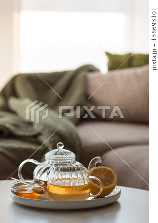 Glass teapot with amber tea, teacup with lemon slice, and bowl of honey with golden spoon on white table in cozy living room Glass teapot with amber tea, teacup with lemon slice, and bowl of honey with golden spoon on white table in cozy living room 138693101