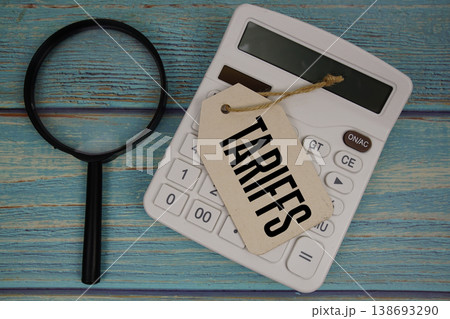 Price tag with "TARIFFS" on calculator and magnifying glass on a rustic blue wooden surface 138693290