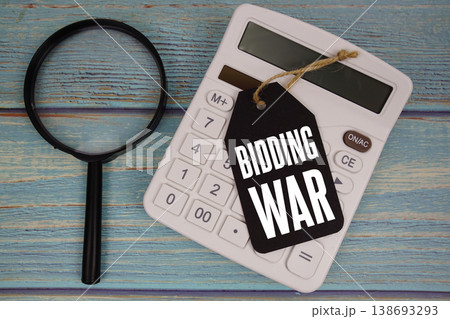 Price tag with "BIDDING WAR" on calculator and magnifying glass on a rustic blue wooden surface 138693293