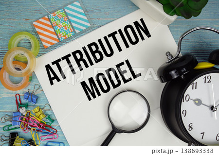 Attribution Model text on a paper surrounded by office supplies, a magnifying glass, and a clock on a blue wooden surface 138693338