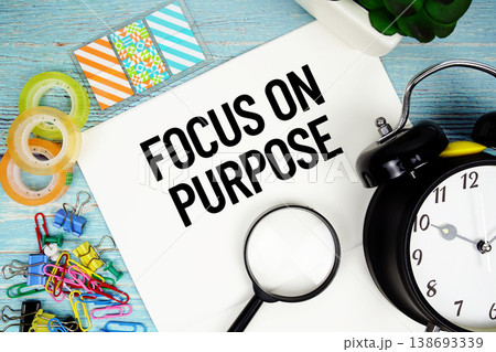 Focus On Purpose text on a paper surrounded by office supplies, a magnifying glass, and a clock on a blue wooden surface Focus On Purpose text on a paper surrounded by office supplies, a magnifying glass, and a clock on a blue wooden surface 138693339