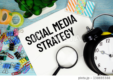 Social Media Strategy text on a paper surrounded by office supplies, a magnifying glass, and a clock on a blue wooden surface 138693388