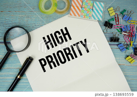 High Priority text on a paper surrounded by office supplies and magnifying glass on a blue wooden surface High Priority text on a paper surrounded by office supplies and magnifying glass on a blue wooden surface 138693559