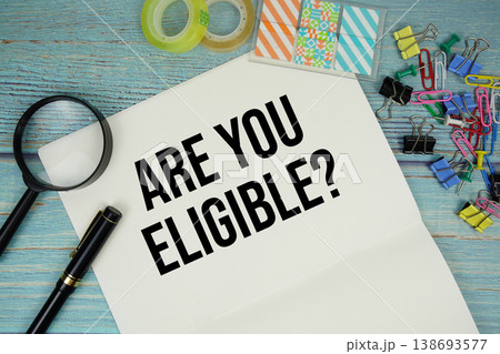 Are You Eligible? text on a paper surrounded by office supplies and magnifying glass on a blue wooden surface 138693577