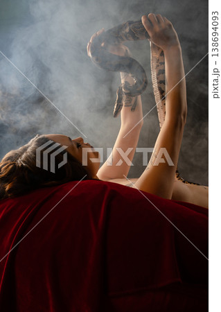 Tattooed woman lying on red velvet holding a large snake in a smoky atmosphere. 138694093