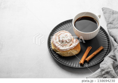 Freshly baked cinnamon roll with white icing and a cup of black coffee on a light background. 138694230