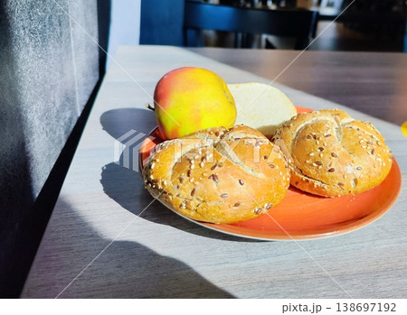 Two kaiser rolls topped with sesame and flax seeds rest on an orange plate. An apple with a red and yellow gradient and a slice of white bread accompany them Two kaiser rolls topped with sesame and flax seeds rest on an orange plate. An apple with a red and yellow gradient and a slice of white bread accompany them 138697192