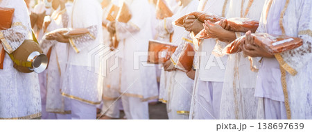 Participants in white clothing line up holding folded orange robes as soft sunlight reveals intricate lace details and hands within a medium to large ceremonial setting. Participants in white clothing line up holding folded orange robes as soft sunlight reveals intricate lace details and hands within a medium to large ceremonial setting. 138697639