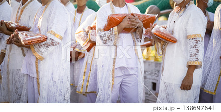 Participants in white clothing line up holding folded orange robes as soft sunlight reveals intricate lace details and hands within a medium to large ceremonial setting. 138697641
