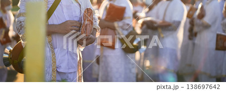Participants in white clothing line up holding folded orange robes as soft sunlight reveals intricate lace details and hands within a medium to large ceremonial setting. 138697642