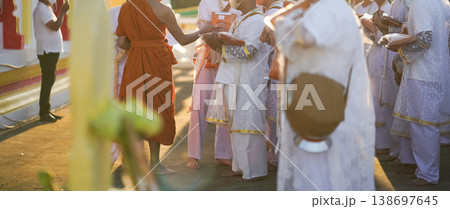 Participants in white clothing line up holding folded orange robes as soft sunlight reveals intricate lace details and hands within a medium to large ceremonial setting. 138697645