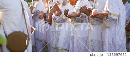 Participants in white clothing line up holding folded orange robes as soft sunlight reveals intricate lace details and hands within a medium to large ceremonial setting. Participants in white clothing line up holding folded orange robes as soft sunlight reveals intricate lace details and hands within a medium to large ceremonial setting. 138697652
