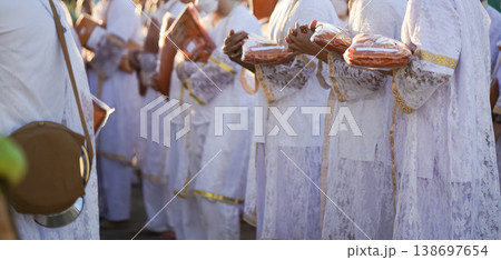Participants in white clothing line up holding folded orange robes as soft sunlight reveals intricate lace details and hands within a medium to large ceremonial setting. Participants in white clothing line up holding folded orange robes as soft sunlight reveals intricate lace details and hands within a medium to large ceremonial setting. 138697654