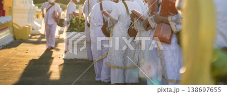 Participants in white clothing line up holding folded orange robes as soft sunlight reveals intricate lace details and hands within a medium to large ceremonial setting. Participants in white clothing line up holding folded orange robes as soft sunlight reveals intricate lace details and hands within a medium to large ceremonial setting. 138697655