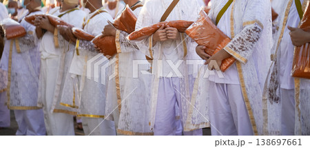 Participants in white clothing line up holding folded orange robes as soft sunlight reveals intricate lace details and hands within a medium to large ceremonial setting. 138697661