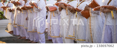 Participants in white clothing line up holding folded orange robes as soft sunlight reveals intricate lace details and hands within a medium to large ceremonial setting. Participants in white clothing line up holding folded orange robes as soft sunlight reveals intricate lace details and hands within a medium to large ceremonial setting. 138697662