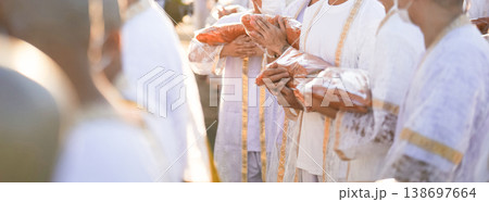 Participants in white clothing line up holding folded orange robes as soft sunlight reveals intricate lace details and hands within a medium to large ceremonial setting. Participants in white clothing line up holding folded orange robes as soft sunlight reveals intricate lace details and hands within a medium to large ceremonial setting. 138697664