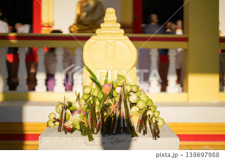 A temple marker stands above arranged lotus incense and candles as fresh lotus bundles and folded robes rest nearby with a blurred statue adding depth to the warm sacred ceremonial setting. 138697988