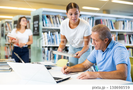 Positive young lady explaining information and pointing finger in a book for mature man while he is using a computer in the library 138698559