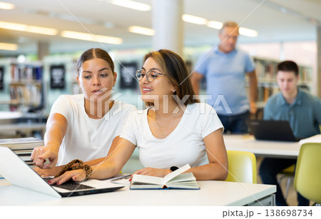Positive female students using laptop and talking with each other in the book space library 138698734