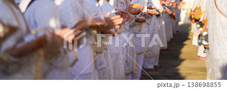 Thai men in white walk in line with palms together carrying folded orange monk robes as medium to large group approaches temple doorway, warm light before ordination. 138698855