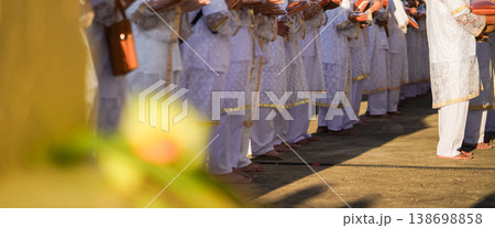 Thai men in white walk in line with palms together carrying folded orange monk robes as medium to large group approaches temple doorway, warm light before ordination. Thai men in white walk in line with palms together carrying folded orange monk robes as medium to large group approaches temple doorway, warm light before ordination. 138698858