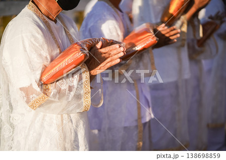 Thai men in white walk in line with palms together carrying folded orange monk robes as medium to large group approaches temple doorway, warm light before ordination. 138698859