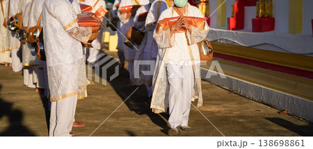 Thai men in white walk in line with palms together carrying folded orange monk robes as medium to large group approaches temple doorway, warm light before ordination. 138698861