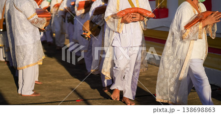 Thai men in white walk in line with palms together carrying folded orange monk robes as medium to large group approaches temple doorway, warm light before ordination. Thai men in white walk in line with palms together carrying folded orange monk robes as medium to large group approaches temple doorway, warm light before ordination. 138698863