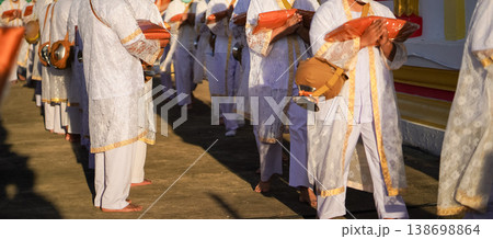 Thai men in white walk in line with palms together carrying folded orange monk robes as medium to large group approaches temple doorway, warm light before ordination. Thai men in white walk in line with palms together carrying folded orange monk robes as medium to large group approaches temple doorway, warm light before ordination. 138698864