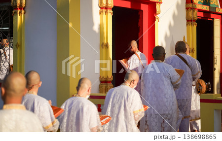 Thai men in white walk in line with palms together carrying folded orange monk robes as medium to large group approaches temple doorway, warm light before ordination. Thai men in white walk in line with palms together carrying folded orange monk robes as medium to large group approaches temple doorway, warm light before ordination. 138698865