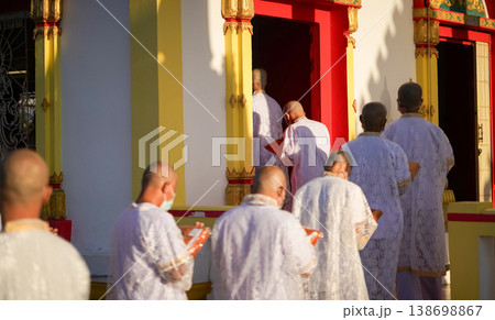 Thai men in white walk in line with palms together carrying folded orange monk robes as medium to large group approaches temple doorway, warm light before ordination. 138698867
