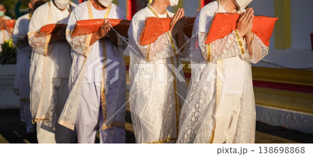 Thai men in white walk in line with palms together carrying folded orange monk robes as medium to large group approaches temple doorway, warm light before ordination. 138698868