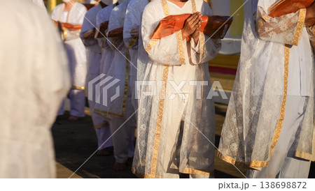 Thai men in white walk in line with palms together carrying folded orange monk robes as medium to large group approaches temple doorway, warm light before ordination. 138698872