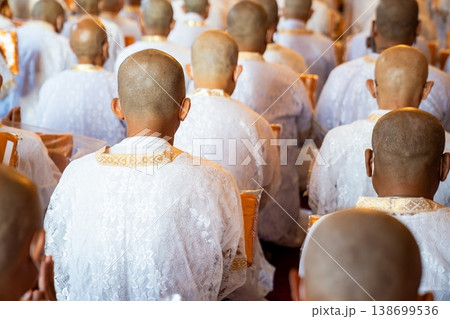 A rear perspective shows white clad ordinands seated in rows inside the temple holding orange robes with shaved heads and folded hands in a soft blurred ritual setting. 138699536