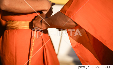 A senior monk adjusts a novice robe as hands tighten the woven belt around orange cloth in sunlight with focus on texture folds and precise fitting. 138699544