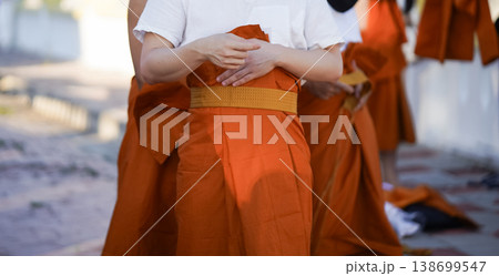 A senior monk adjusts a novice robe as hands tighten the woven belt around orange cloth in sunlight with focus on texture folds and precise fitting. 138699547