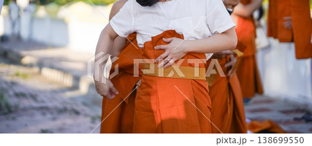 A senior monk adjusts a novice robe as hands tighten the woven belt around orange cloth in sunlight with focus on texture folds and precise fitting. 138699550