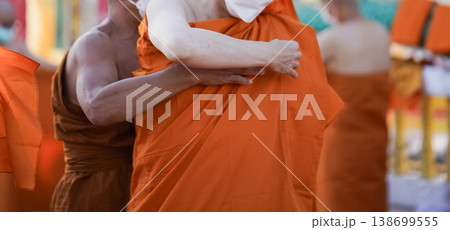 A senior monk adjusts a novice robe as hands tighten the woven belt around orange cloth in sunlight with focus on texture folds and precise fitting. 138699555
