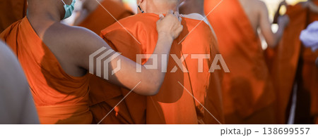 A senior monk adjusts a novice robe as hands tighten the woven belt around orange cloth in sunlight with focus on texture folds and precise fitting. 138699557