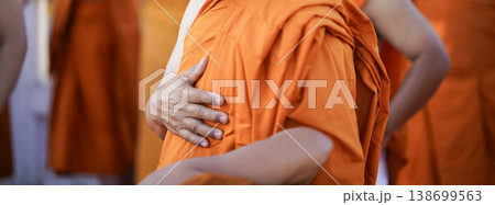 A senior monk adjusts a novice robe as hands tighten the woven belt around orange cloth in sunlight with focus on texture folds and precise fitting. 138699563