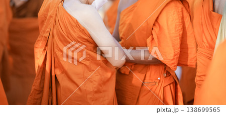 A senior monk adjusts a novice robe as hands tighten the woven belt around orange cloth in sunlight with focus on texture folds and precise fitting. A senior monk adjusts a novice robe as hands tighten the woven belt around orange cloth in sunlight with focus on texture folds and precise fitting. 138699565